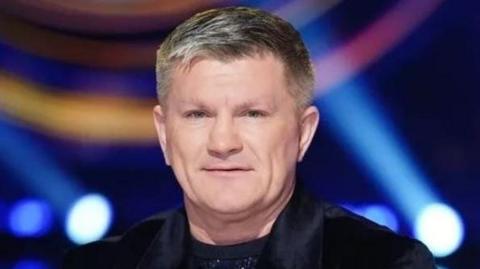 Ricky Hatton with light brown hair wearing a black velvet jacket and dark crew-neck top with spotlights shining behind him. He is smiling.