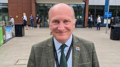 Nick Hulme is standing in front of Colchester hospital  in green checked jacket with rainbow pin and “Chief Executive” lanyard. He is smiling at the camera. 
