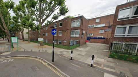 A Street View image showing Courtauld Road, on the corner of Beachcroft Way, with a small lamppost and two bollards in front of small blocks of flats