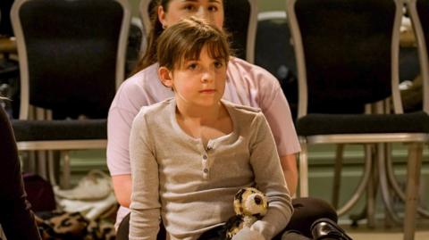 Nine-year-old Evie sitting on the floor holding a soft toy of an owl, there is an older girl sitting behind her wearing a light pink T-shirt