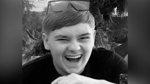 A black and white picture of the teenager who is laughing. He wears sunglasses on the top of his head, trees and plants can be seen in the background.