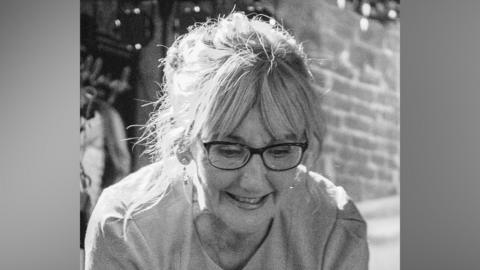 A black-and-white photograph of Susan Watson. She has blonde hair and is wearing glasses. She is leaning forward.