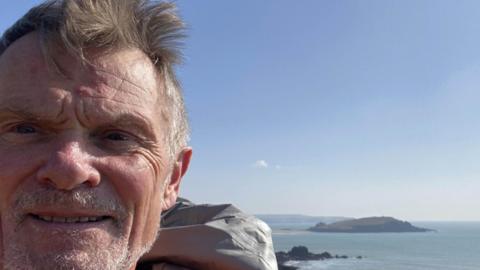A man smiling at a camera. He has a beard and is stood in front of a body of water with an island in the background.