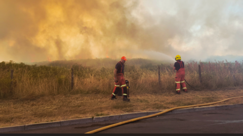 Two firefighters, one using a hose to spray water over a burning field. A haze of deep yellow smoke hangs over an area of dry, brown grass.The two men are wearing red, high-vis overalls, one wears a yellow protective helmet, and the other wears a red baseball cap.