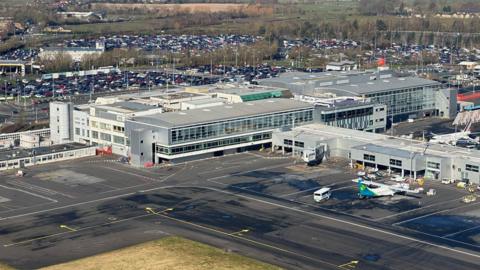 Aerial view of Newcastle Airport. There is a large terminal building and several planes parked outside, with a large car park beyond