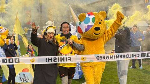 Pudsey bear, a yellow bear with a spotty eyepatch alongside Cate Conway, who has light hair. She is wearing yellow bear ears and a black, she is beside Conor Phillips who has dark hair. He is wearing a yellow top and holding a microphone. All three are at a BBC finish line surrounding by on lookers, flags and yellow smoke from flares.
