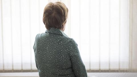 Stock image of a woman with short hair wearing a grey/green dressing gown with white spots on it, looking out towards a window covered in vertical blinds