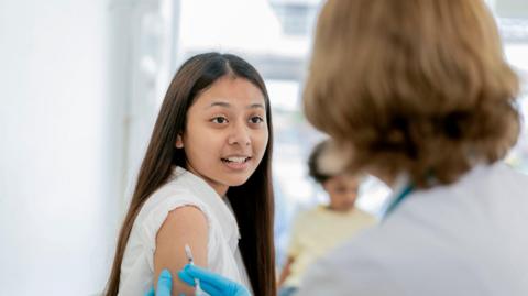 Stock image of young lady having a vaccination. She has dark long hair and wears a white top. She is looking at a blurred person in the foreground who is administering an injection to the lady's upper arm. The healthcare professional is wearing blue gloves. 