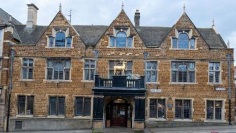 Historic stone building with ornate architecture, featuring multiple pointed gables, large mullioned windows, and a central entrance with a black balcony topped by a golden hind statue. The building has signs reading 'The Hind Hotel' and is situated along a paved street with two red bollards in the foreground.