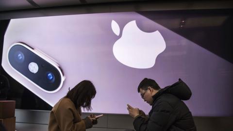 Two people look at their iPhones in an Apple store, dwarfed by an enormous promotional image for the latest model