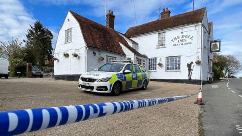 A pub in Colchester. The building is white and has The Bell Inn Gray & Sons written on it. A Essex Police car is parked at the pub and a police cordon has been put in place