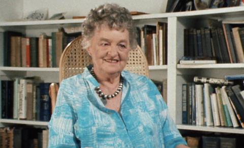 P. L. Travers sitting on a wicker chair in front of bookshelves, wearing a blue patterned shirt.