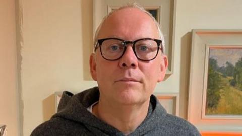 Gary Temprell is wearing a grey hooded top and dark rimmed spectacles. Behind him are a number of pictures on the wall of his home.
