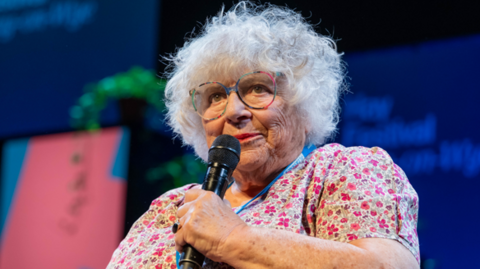 Close up of Margoyles talking on stage at Hay Festival. She wears a floral dress, colourful glasses and has short curly grey hair. She holds a black microphone and looks out at audience.