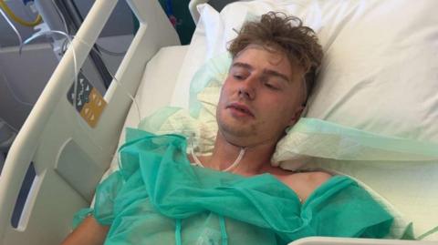 A man lying in a hospital bed. He is attached to tubes and wearing a green gown.