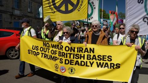 Protesters at the front of a demonstration are holding a large yellow anti-war banner, with many people following behind them.