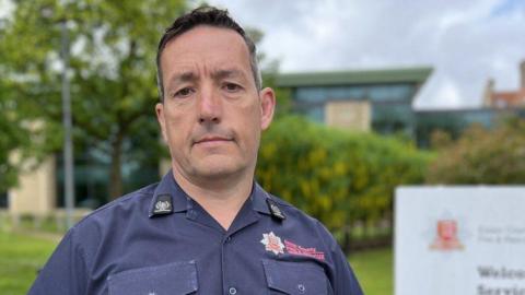 Craig McLellan, who has short black hair, standing in front of a green bush at Essex fire service's headquarters. He looks serious and is wearing a navy shirt.