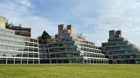 Ziggurat buildings at the University of East Anglia. There are six stories of windows, with three buildings shown. Green grass can be seen at the bottom of the image, and above the buildings the sky is blue with a few wispy clouds. 