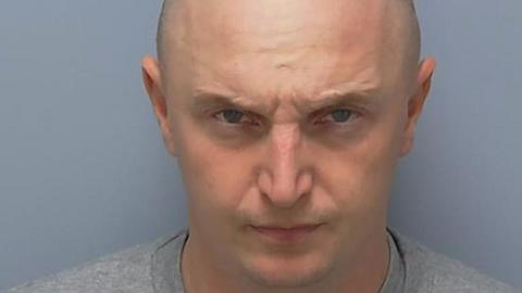 Adam Wright frowns at the camera in a police custody photo. His head is shaved and he wears a grey, collarless top.