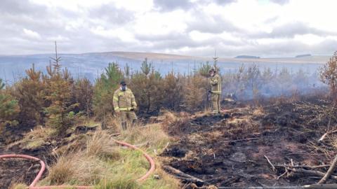 Two firefighters with a large hose on Langdale Moor. They are facing a burned out patch of ground. There is smoke hanging in the air around them