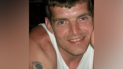 A man in a white vest and with a small Chinese taijitu tattoo on his right shoulder smiling at the camera