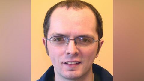 A man is standing in front of a yellow wall. He has dark, receding hair and glasses. He is wearing a green t-shirt and navy fleece.
