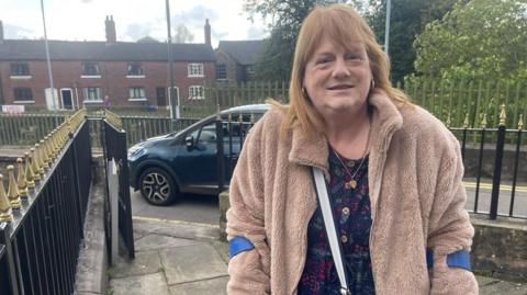 Karen Bradley has auburn hair past her shoulders and wears a light brown fleece top over a dress while having the tops of crutches visible around her arms. She is stood on paving inside iron fencing