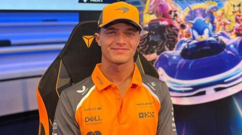 Lando Norris wearing an orange cap, and an orange polo shirt. He is sitting in front of a poster which includes sonic the hedgehog in a blue car.