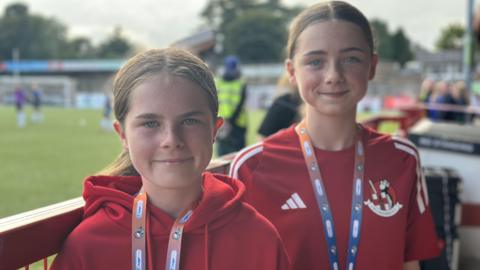 Ami and Lexi have red tops on and SuperCup NI lanyards. Both have their brown hair tied back in a ponytail and are standing at the seats by the football pitch. 