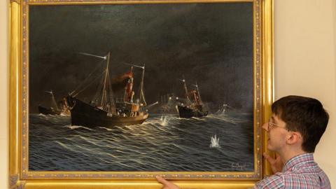 A man with glasses and a stripy shirt is holding up a gold frame on to a wall that shows a dark and stormy painting of boats upon the water. It depicts the Dogger Bank Incident.