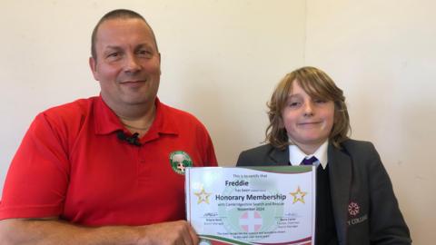 Wayne Bent wearing a red polo top, smiling. He is seen holding a certificate next to Freddie, who has brown, shoulder-length hair and is wearing a white shirt, tie, jumper and blazer.