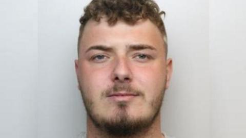 A police custody image of a man with short brown hair and a brown close-trimmed beard.