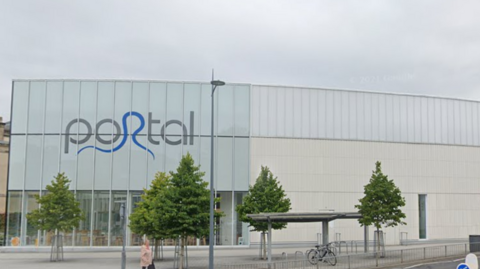 A large leisure centre building, with PORTAL written in grey and blue letters. Several trees sit on the street outside the building.  