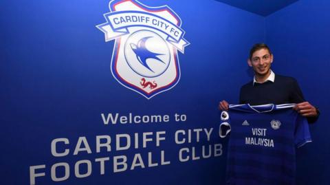 A young man with brown hair, wearing a white shirt and dark jumper. He is holding a blue Cardiff City football shirt and is stood in front of a blue, Cardiff City branded wall which reads "Welcome to Cardiff City Football Club". He is smiling and looking at the camera.