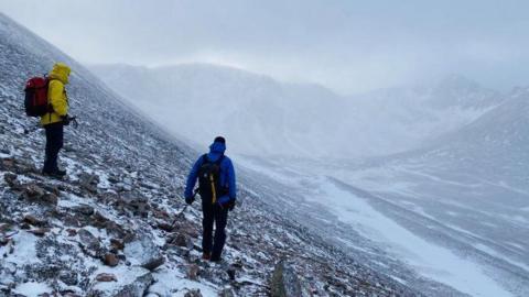 Two walkers, one wearing a yellow walking jacket and other a blue one, stand looking into the coire. There is a dusting of snow.