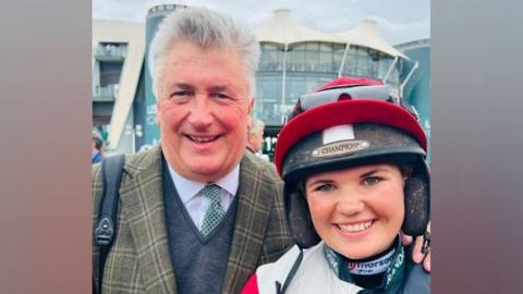 Paul Nicholls and his daughter Olive. Paul is a middle aged man with white hair and is wearing a shirt, tie, v-neck jumper and a tweed jacket. His daughter Olive is wearing a riding helmet with a red cover. 