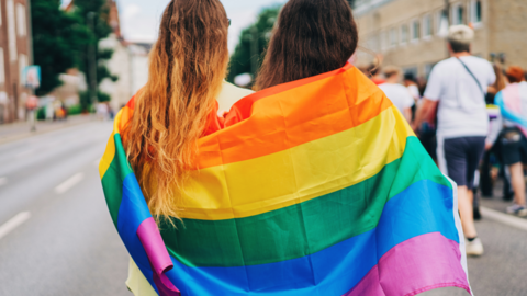 Two women with the rainbow flag wrapped around them, standing on a road behind other people as a parade takes place.