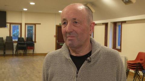 Ruairidh Peutan na sheasamh ann an talla