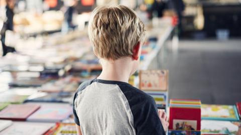 A stock image of a boy with short, blonde hair looking at a series of books. He has his back to the camera and it is unclear what books he is looking at. 
