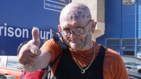 Hoganson gives a thumbs up to the camera as he leaves HMP Durham. He is bald with symbols tattooed all over his head, neck, arm and hand, and is wearing an orange T-shirt with a black vest over the top. Behind him is a sign saying HMP Durham and a car park large blue wall.