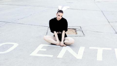 DJ Boo is sat cross-legged next to a white 'No Entry' sign on the floor. She is wearing black shorts and a fluffy black top. On her head is a white headband with bunny ears on.