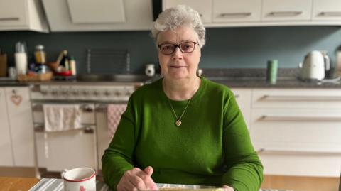 Jennifer is a woman with white hair and black-rimmed spectacles. She is wearing a green wool jumper with a gold heart on a chain round her neck and is sitting in a modern kitchen.