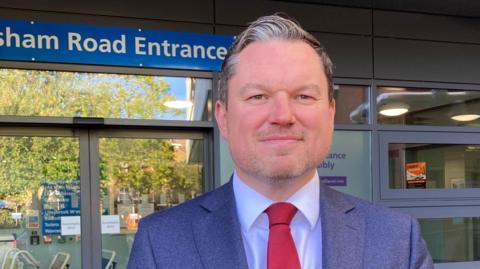 A middle aged man with greying brown hair wears a navy suit with a navy shirt and red tie as he stands outside a hospital entrance. He is slightly smiling. Wheelchairs can be seen in a line on the other side of the doors, inside the hospital.