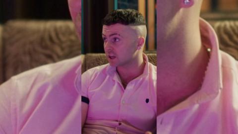 Conor has short dark hair with a skin-fade, and is wearing a pink polo-shirt.