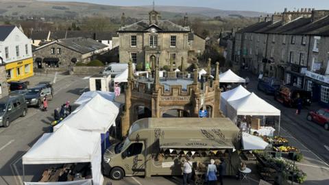 The town square in Kirkby Lonsdale is bordered by stone buildings, the one at the far end has tall arched windows and a clock above it. There is a pop-up market in the centre with white tents erected and a dark green food van. There is a stone structure in the centre with a stone cross atop it.