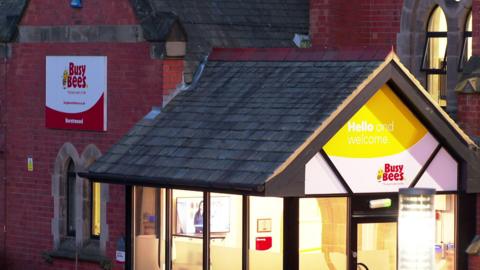 A large red brick building with a sloped roof and arched windows. We can see a 'Hello and welcome to Busy Bees' sign. The lights are on bright. It is evening time so the rest of the building is slightly dark. 