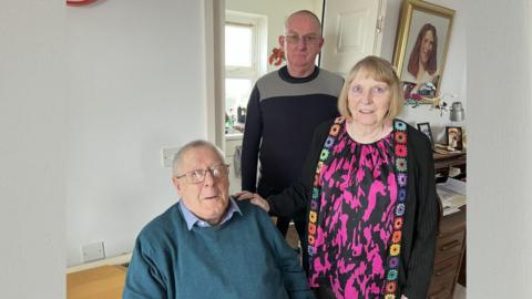 Three people are pictured inside of a flat. One person is sat down wearing a blue sweater. Another person, a woman in her 80s, is standing up. She is in front of another man, wearing a black and grey jumper.