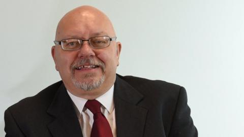 Glyn Jones looks at the camera. He is bald and has a greying goatee beard.  He is wearing rectangular shaped glasses.  He is wearing a black blazer, white shirt and burgundy coloured tie and is smiling.