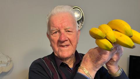 Wally Newman is 100 years old and has short grey hair swept back, he is holding a bunch of bananas on the right, he is wearing a dark/purple woollen jumper with a purlpe shirt and blue t-shirt below, behind him is a living room magnolia wall, with part of a lampshade, tv and clock visible.