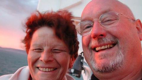 A happy couple smile as they cuddle up together. It is sunset by the seaside. He is bald with a goatee beard and glasses. She had short red hair.